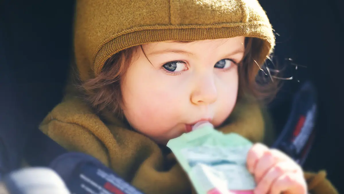 Close up portrait of sweet toddler kid eating fruit puree from plastic doy pack, sitting in stroller, outdoor snack time