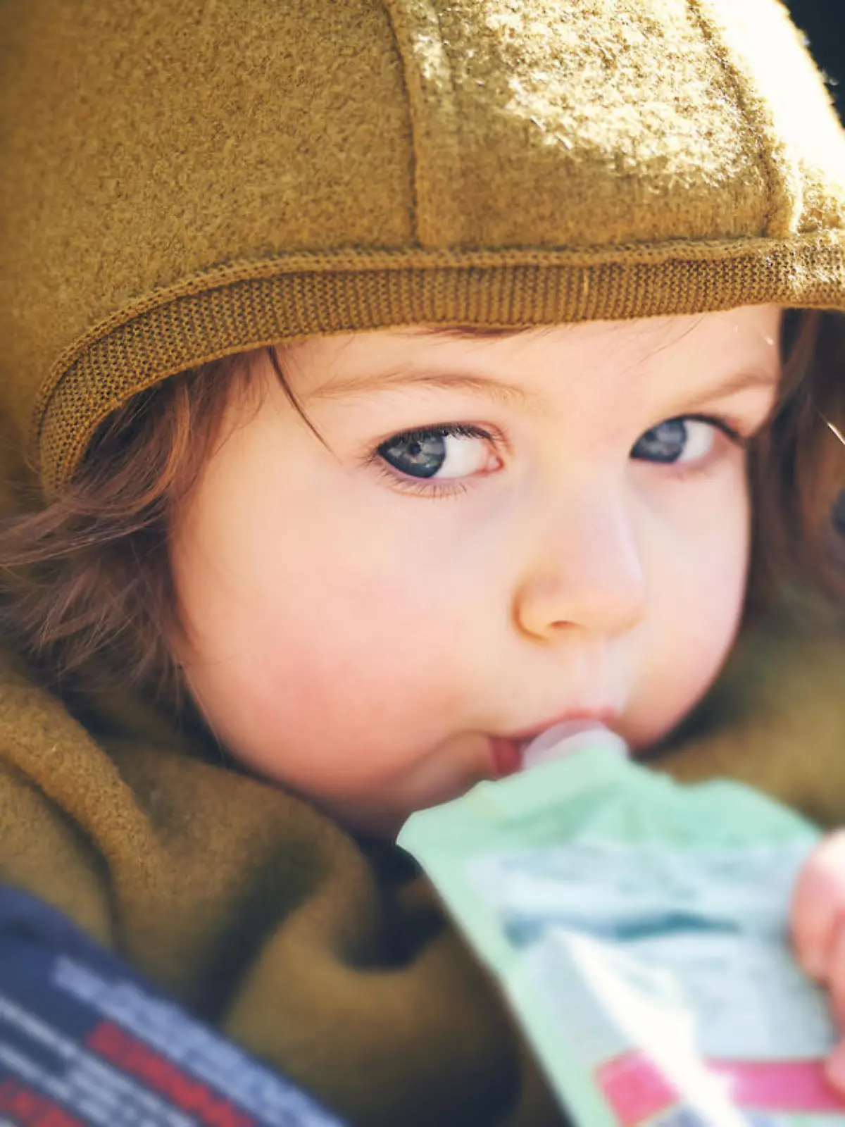 Close up portrait of sweet toddler kid eating fruit puree from plastic doy pack, sitting in stroller, outdoor snack time