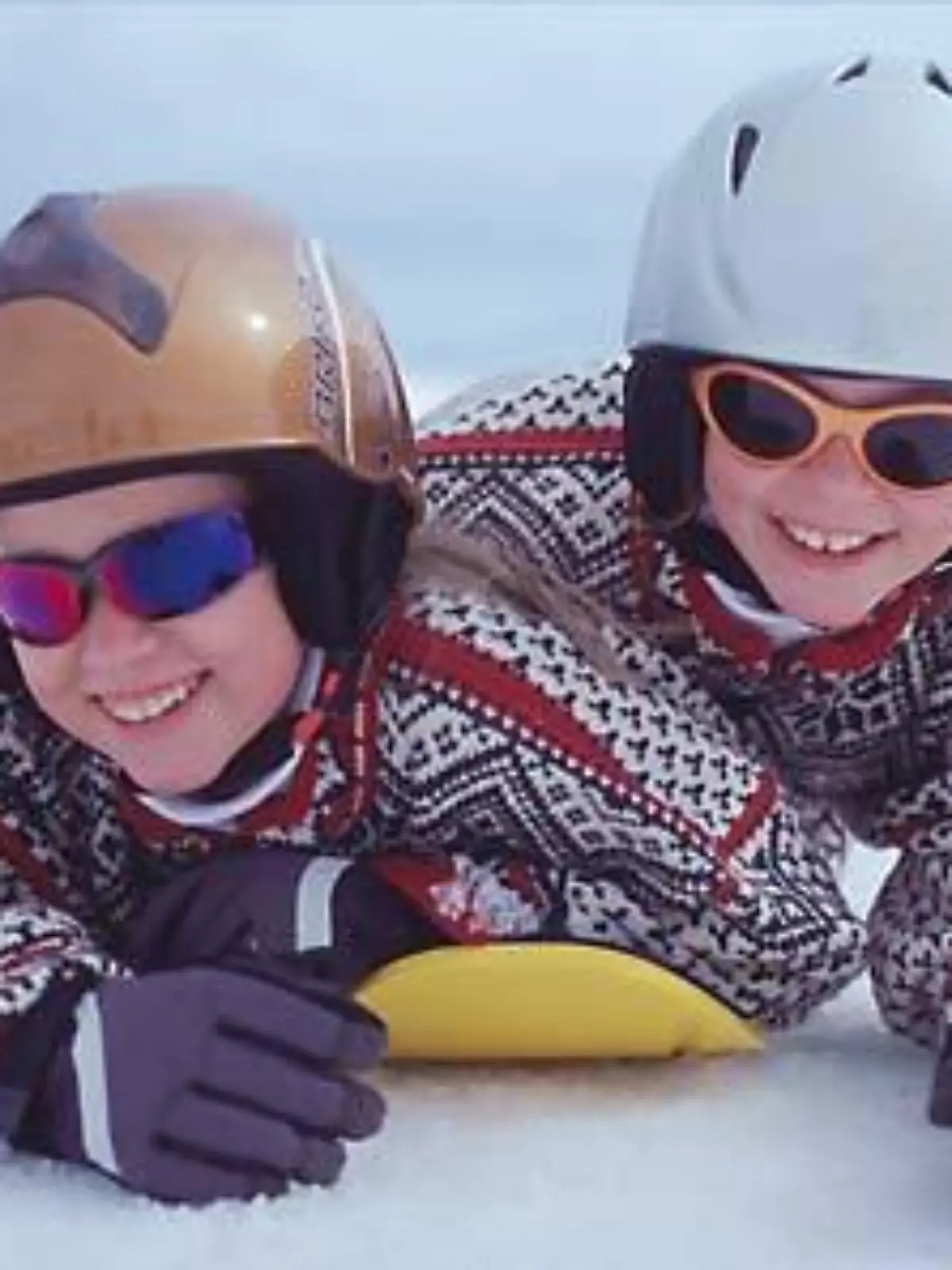 Zwei Kinder liegen auf Mini-Snowboard mit Helmen.