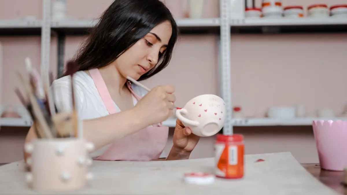 Beautiful young woman is painting a ceramic mug in workshop