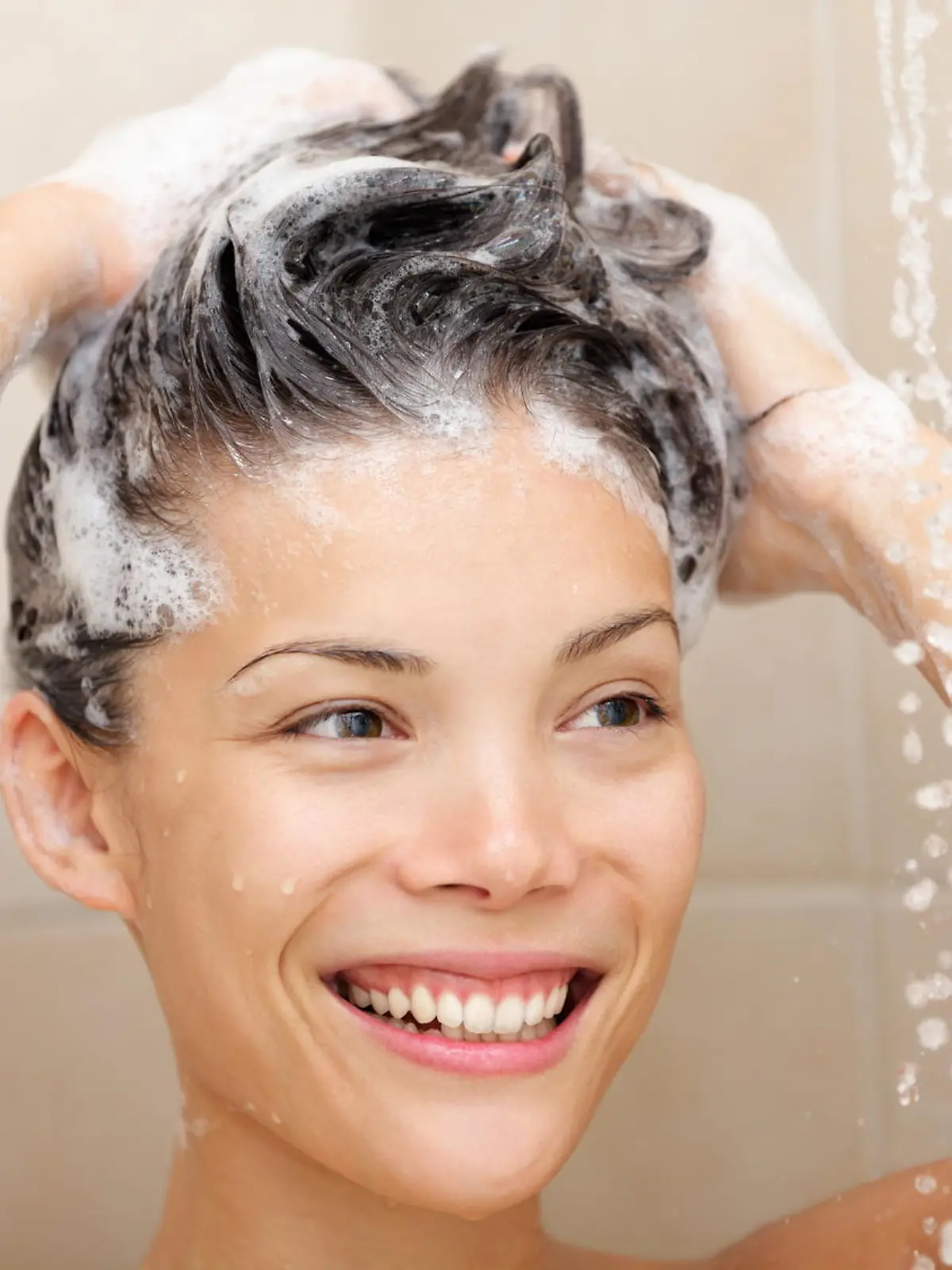 Woman washing hair with shampoo foam in shower smiling happy looking at running warm water. Beautiful mixed race Asian Chinese / Caucasian female model in bathroom.