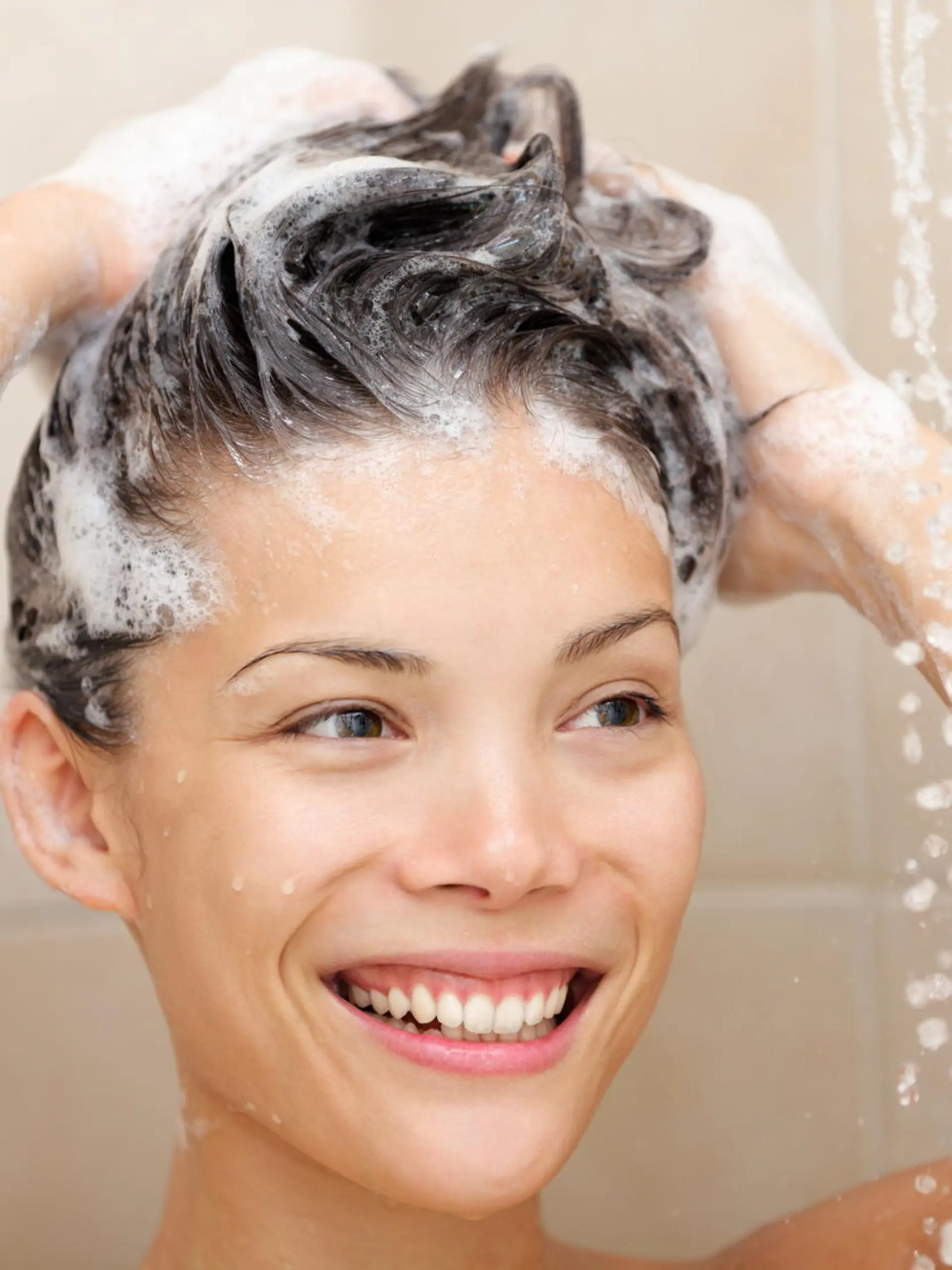 Woman washing hair with shampoo foam in shower smiling happy looking at running warm water. Beautiful mixed race Asian Chinese / Caucasian female model in bathroom.