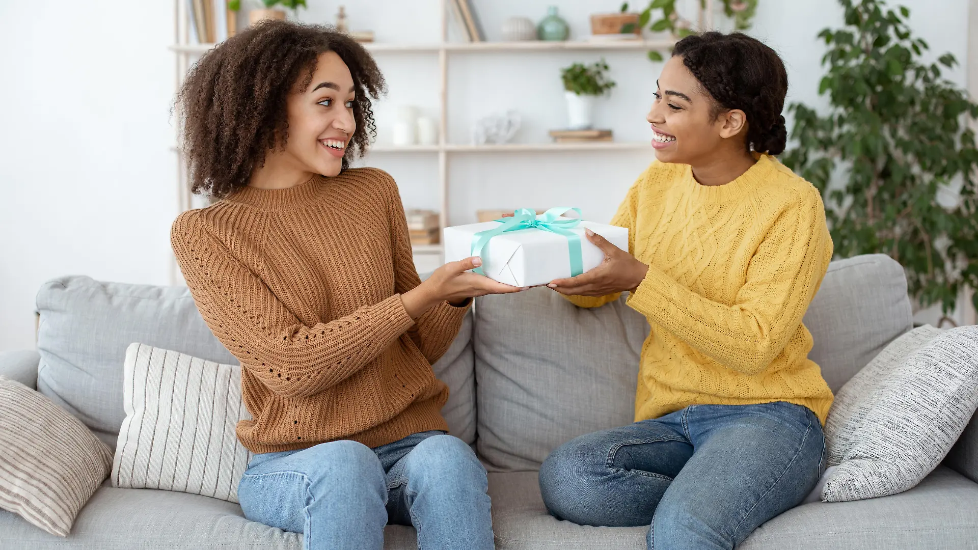 Two ladies friends making surprise birthday present. Happy millennial african american female congratulates, gives gift to embarrassed mixed race sister, sits on sofa in interior of living room