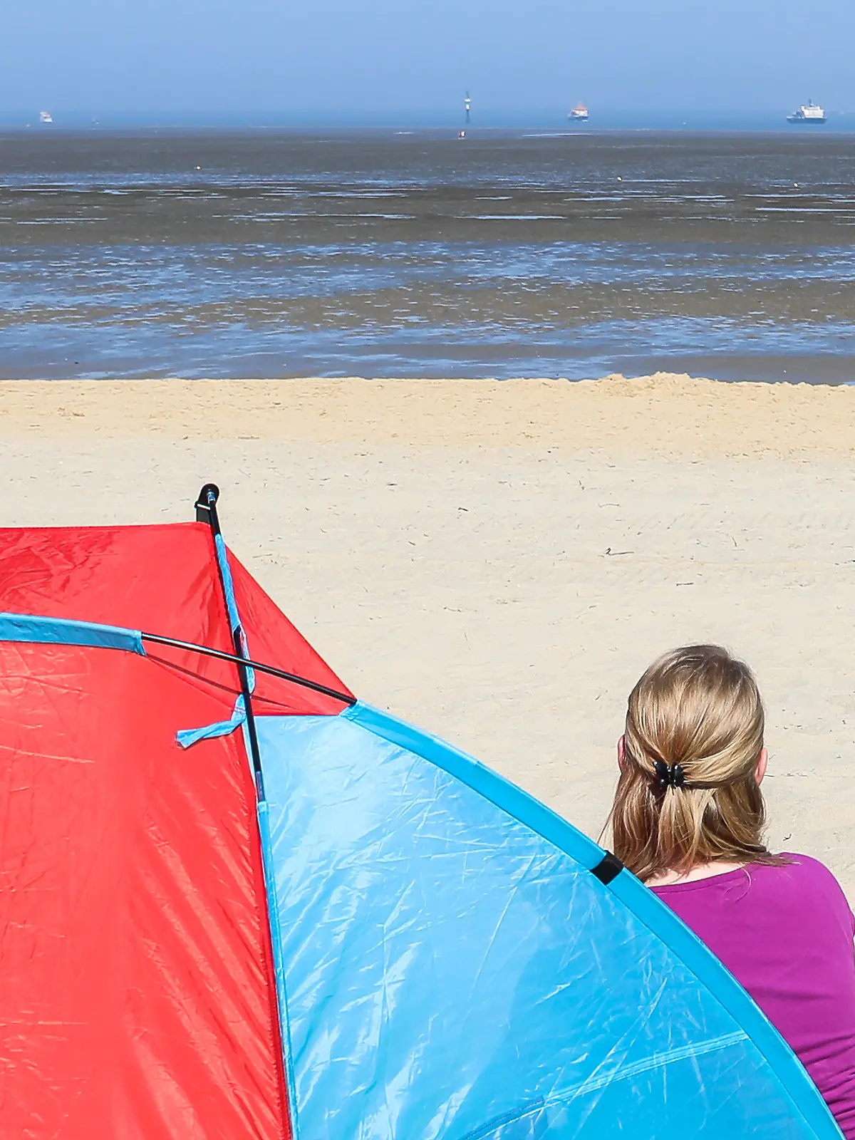 Frau sitzt neben einer rot-blauen Strandmuschel und ist von hinten zu sehen.