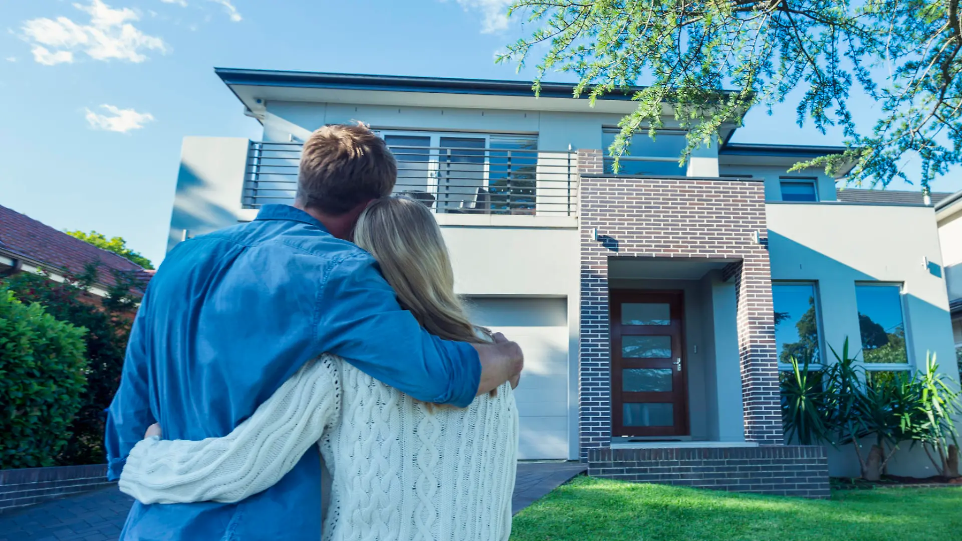 Couple standing in front of their new home. They are both wearing casual clothes and embracing. Rear view from behind them. The house is contemporary with a brick facade, driveway, balcony and a green lawn. The front door is also visible. Copy space