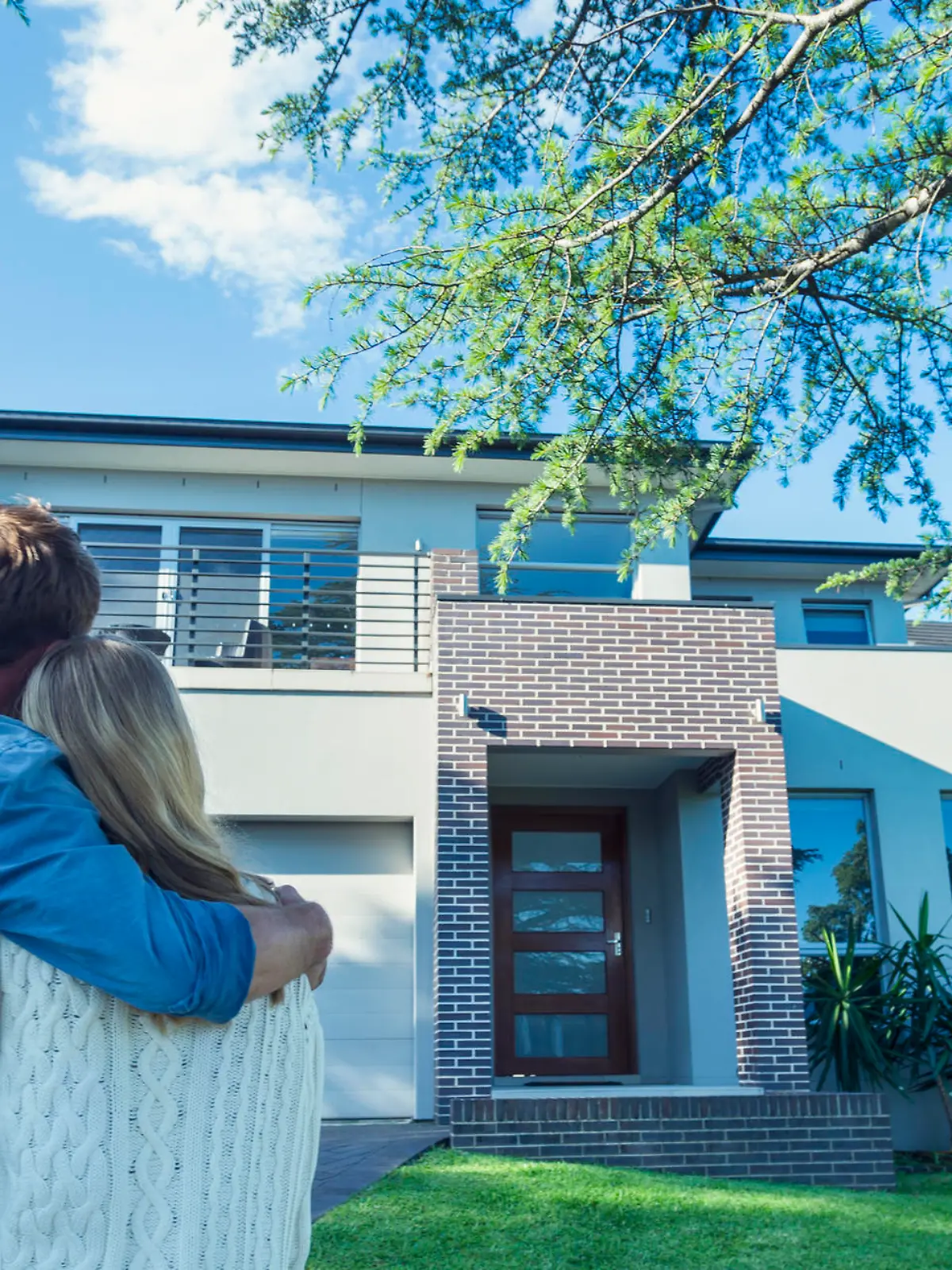 Couple standing in front of their new home. They are both wearing casual clothes and embracing. Rear view from behind them. The house is contemporary with a brick facade, driveway, balcony and a green lawn. The front door is also visible. Copy space