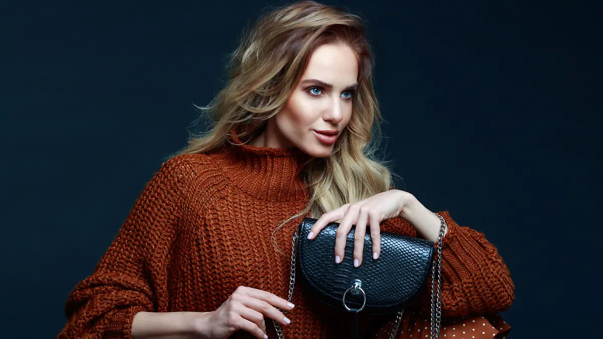 Portrait of long hair blond young woman wearing brown sweater and skirt, holding black purse, looking away. Studio shot against black background.