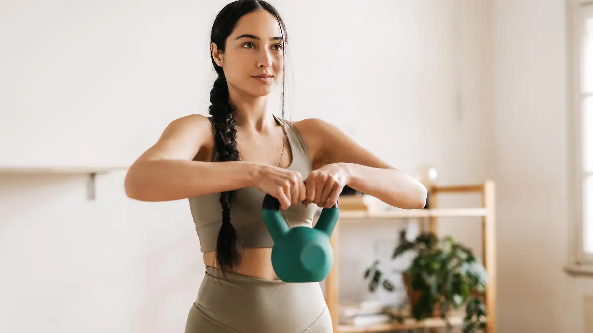 A sporty woman is exercising with a green kettlebell at home. Her eyes are focused. She is dressed in matching fashionable sports leggings and top. Furniture with a plant is visible behind her. Horizontal daylight photo.