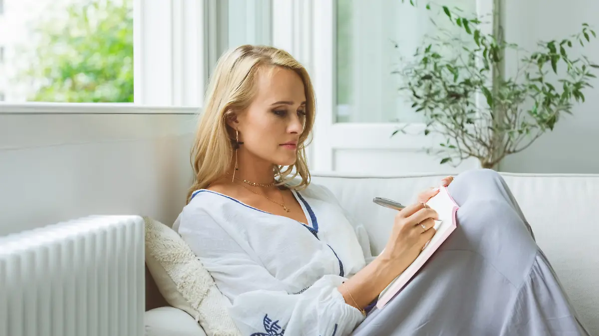 Focused mid adult woman writing in book while sitting on sofa at home
