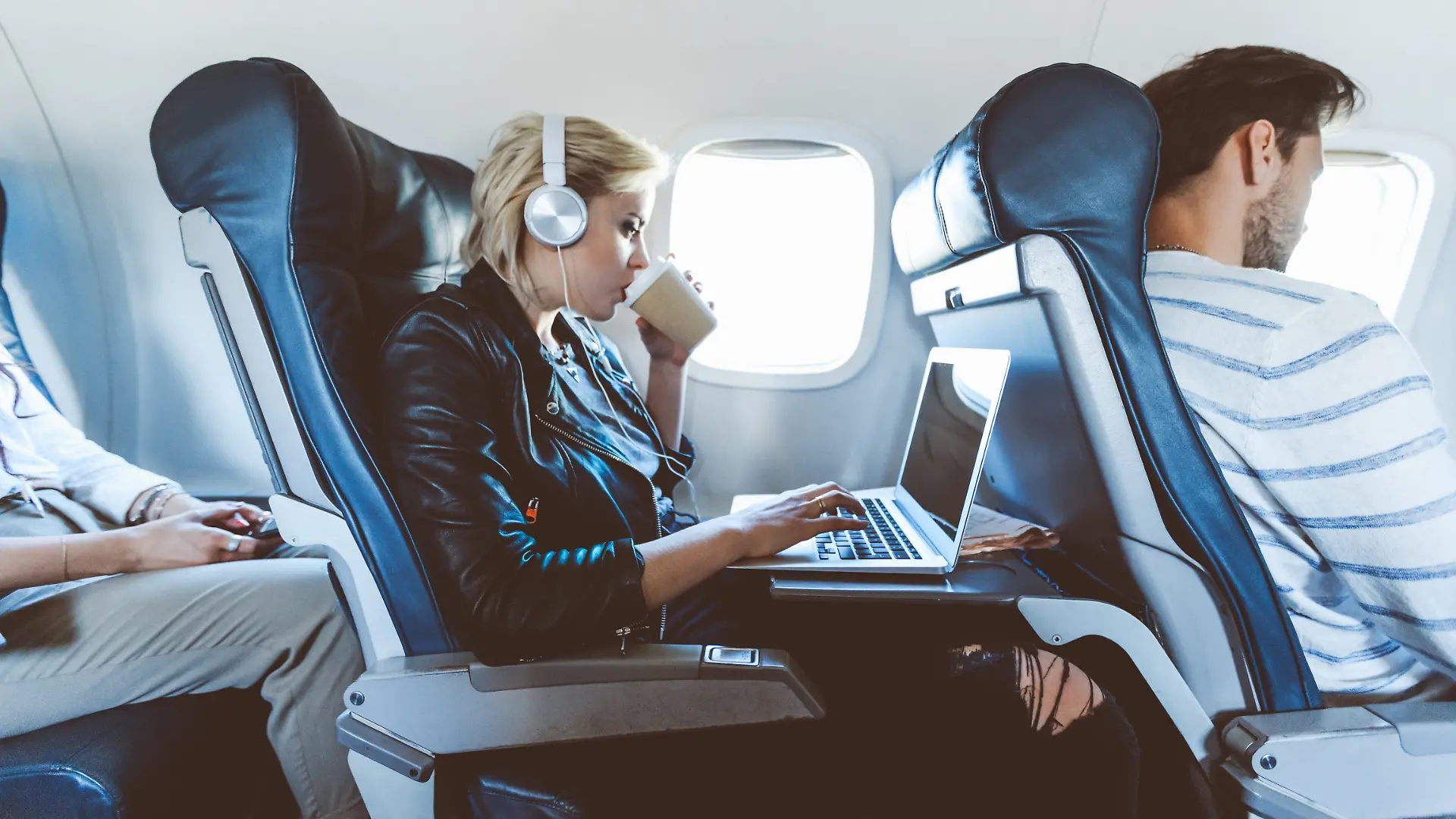 Eine junge blonde Frau sitzt im Flugzeug und arbeitet am Laptop und trinkt dabei Kaffee.