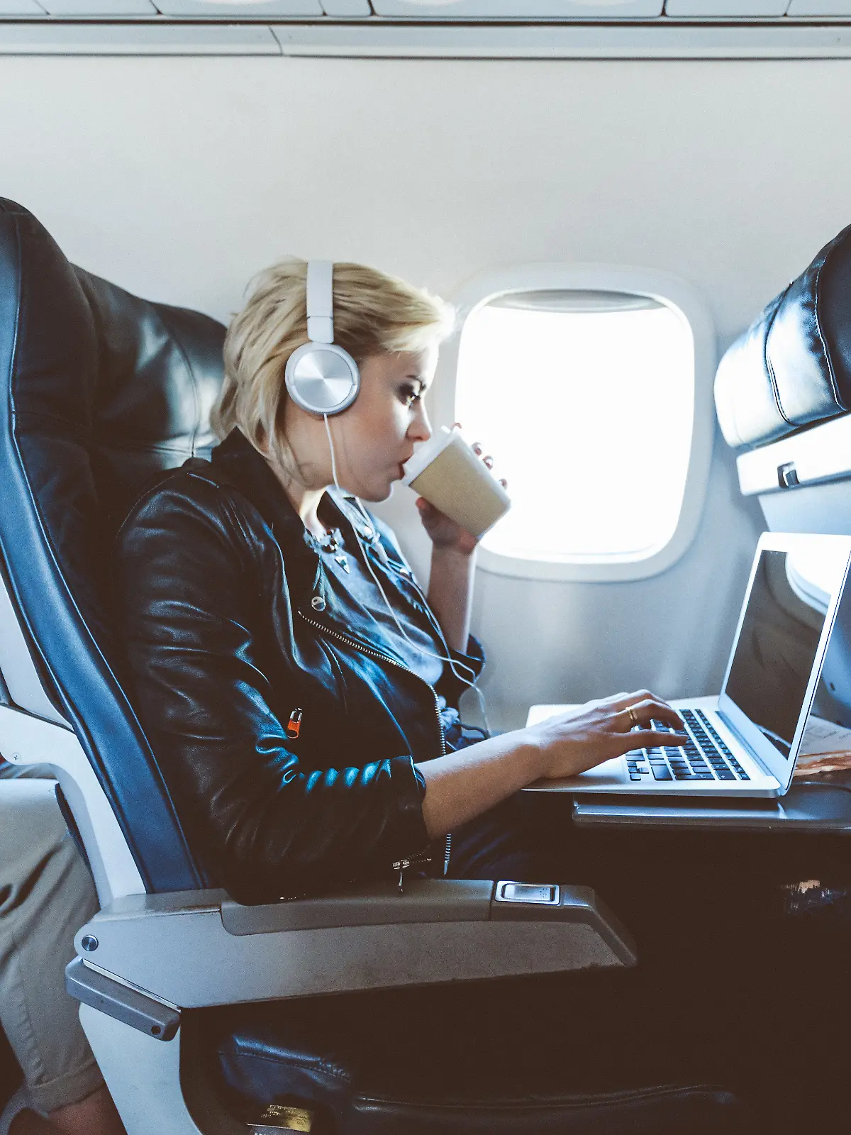 Eine junge blonde Frau sitzt im Flugzeug und arbeitet am Laptop und trinkt dabei Kaffee.