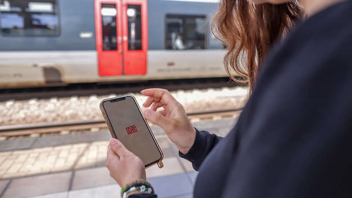 Eine Frau steht mit ihrem Mobiltelefon und der App DB-Navigator auf einem Bahnsteig.