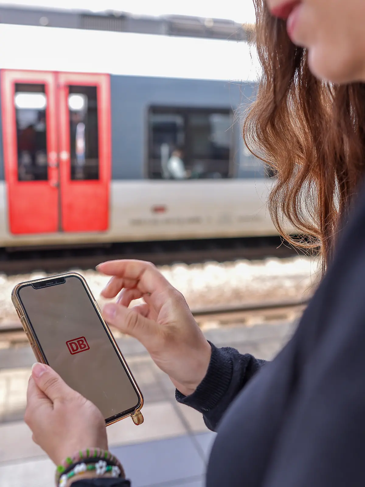 Eine Frau steht mit ihrem Mobiltelefon und der App DB-Navigator auf einem Bahnsteig.
