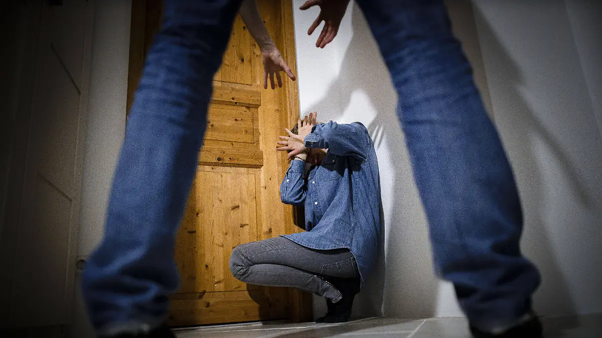 Eine junge Frau sitzt auf dem Boden und hält sich schützend die Hände vor ihr Gesicht. Ein Mann droht, sie anzugreifen (Stockbild).