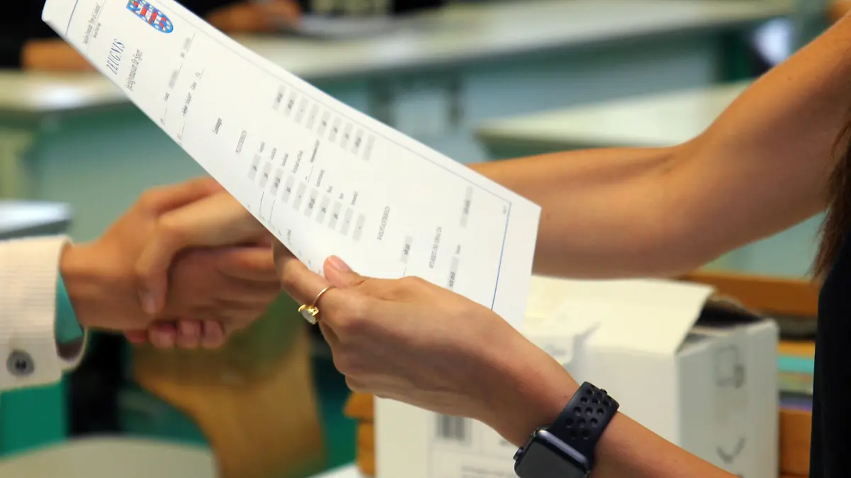 Eine Lehrerin eines Gymnasiums steht am letzten Schultag vor den Sommerferien mit einem Zeugnis in der Hand in einem Klassenzimmer und schüttelt einer Schülerin bei der Zeugnisübergabe die Hand.
