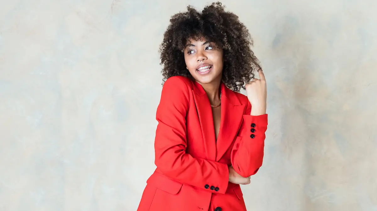 Fashion portrait of curly hair young woman wearing red suit, standing with arms crossed and looking away. Studio shot on beige background.