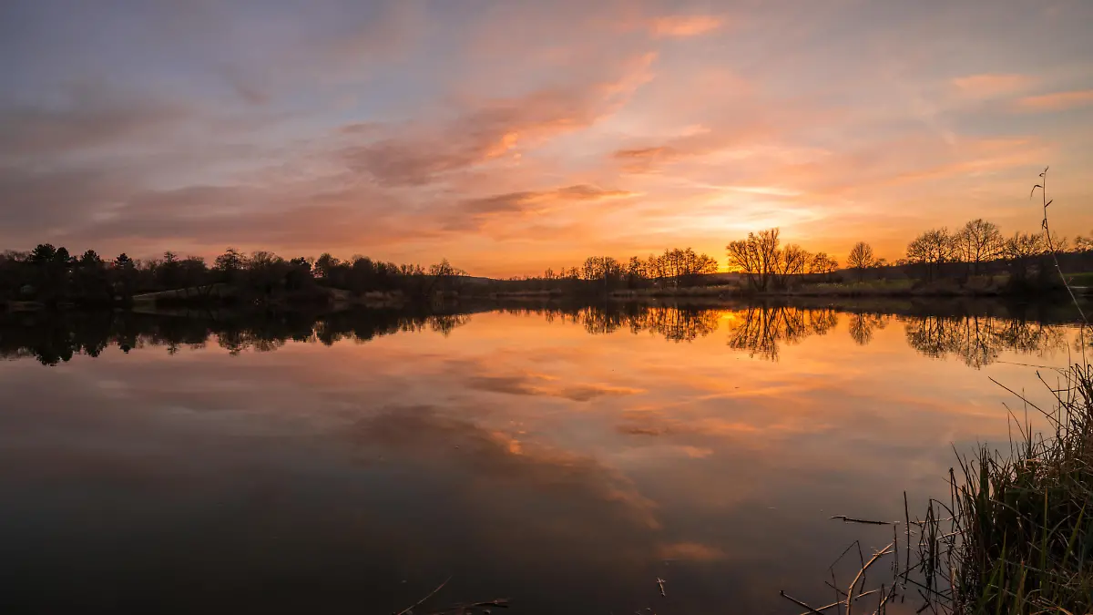 Landscape and sunset on the shore in Westpark Donaupark in Regensburg with reflection of sky in the smooth lake with beautiful dramatic play of colors play with clouds, Germany