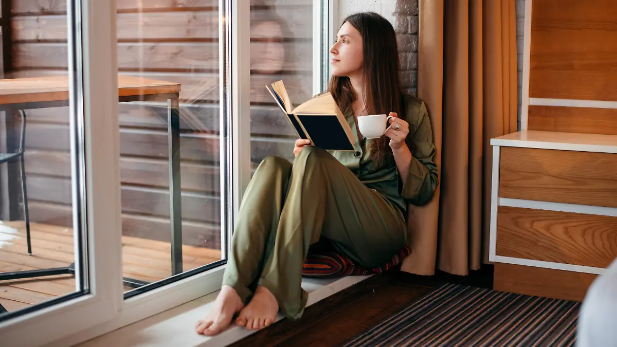 European girl reading book and drinking tea or coffee at home. Young beautiful woman with brown hair wear pajamas. Concept of rest. Modern apartment. Daytime