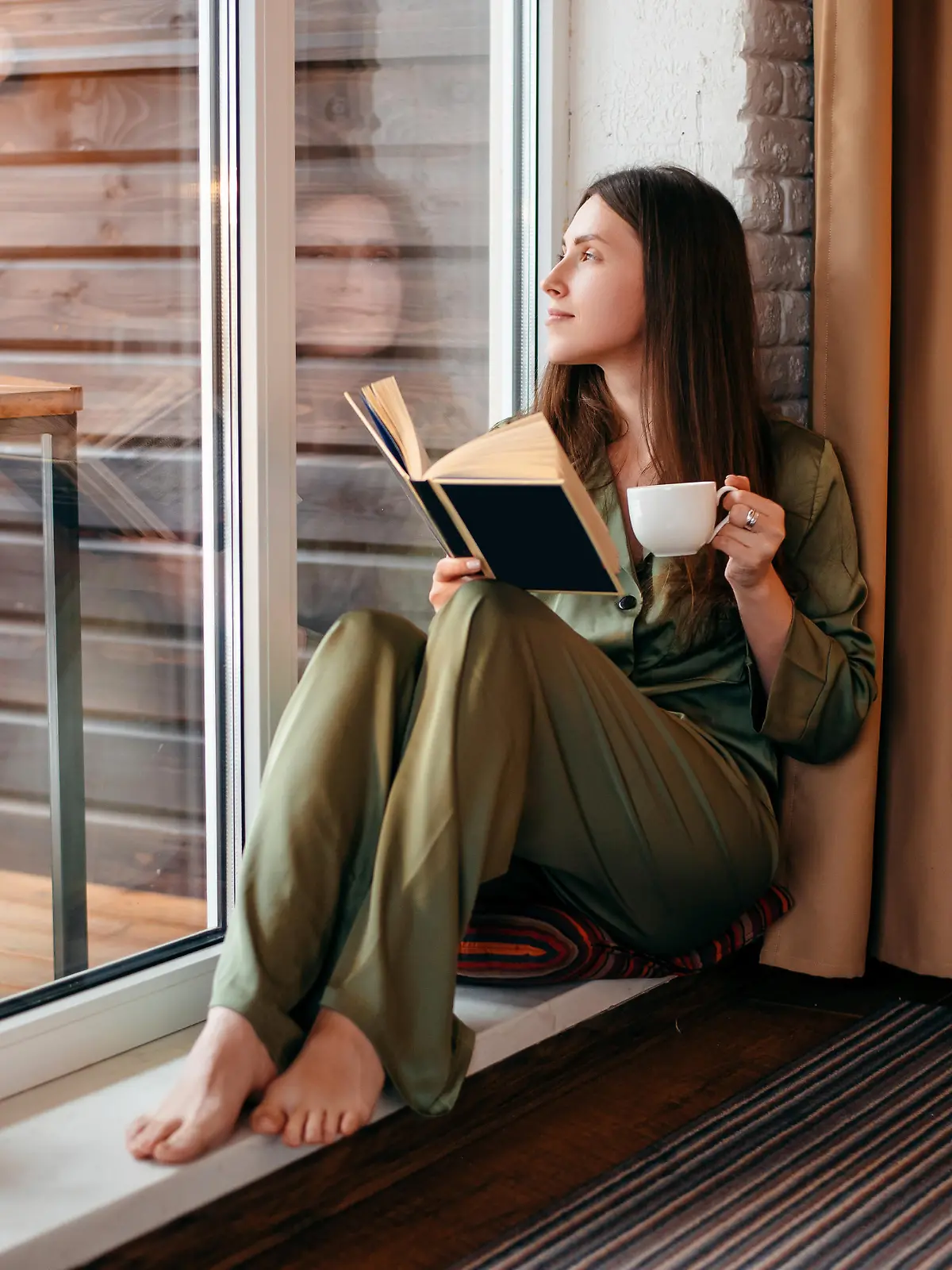 European girl reading book and drinking tea or coffee at home. Young beautiful woman with brown hair wear pajamas. Concept of rest. Modern apartment. Daytime