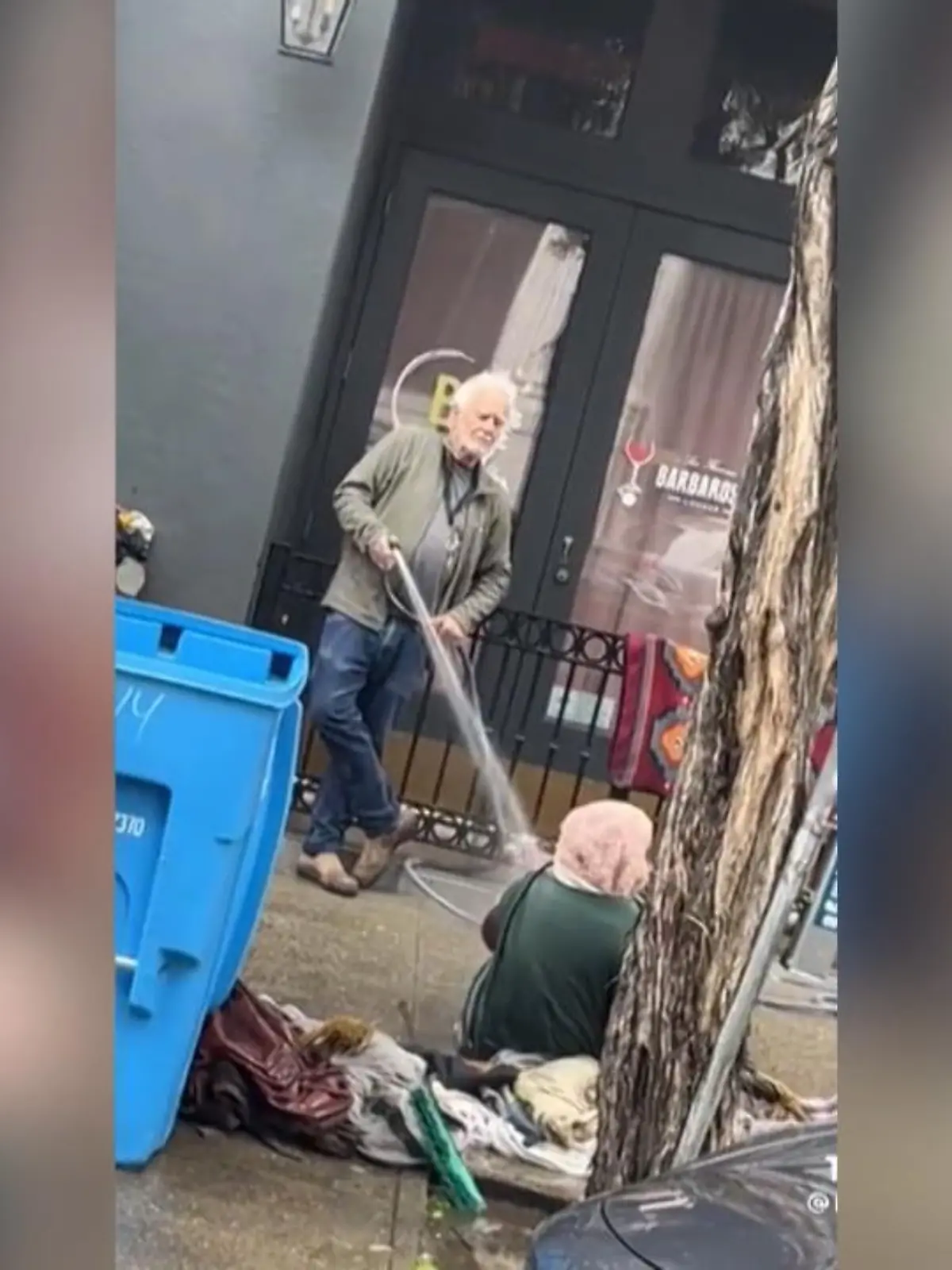 Dieser Galeriebesitzer wollte eine obdachlose Frau vor seinem Laden vertreiben und greift dabei zu drastischen Mitteln.  Während er sich lässig an einen Zaun lehnt, spritzt er die Frau mit einem Gartenschlauch ab.