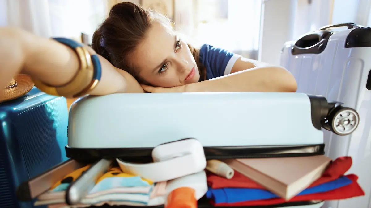 stressed stylish tourist woman in blue t-shirt in the modern house in sunny summer day relaxing on over packed suitcase.