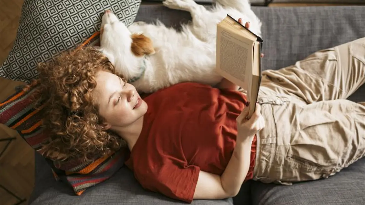 Frau liest ein Buch auf der Couch und kuschelt mit einem Hund.