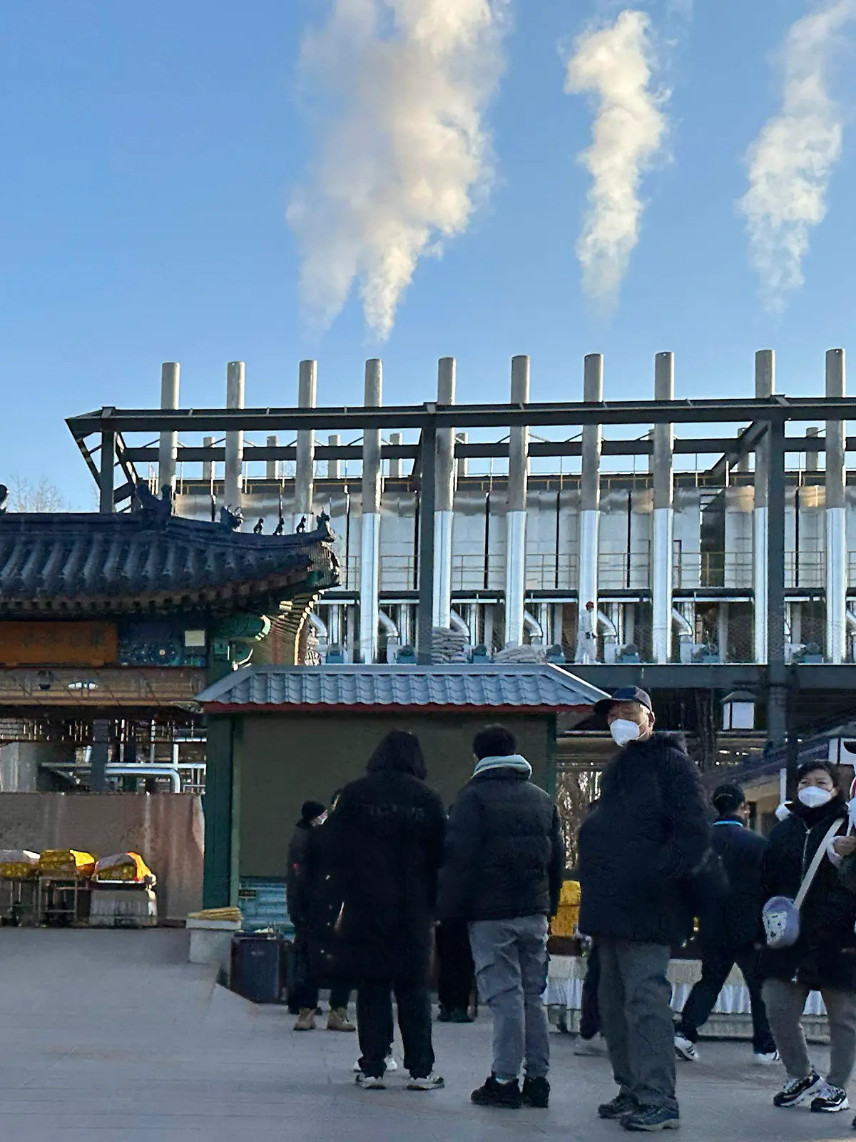31.12.2022, China, Peking: Trauernde stehen vor einem Krematorium, während Rauchsäulen aus den Schornsteinen für die Einäscherung aufsteigen. China hat Anfang dieses Monats viele seiner strengen Corona-Beschränkungen aufgehoben, so dass sich das Virus rasch ausbreiten konnte. Die Zahl der Einäscherungen ist um ein Vielfaches gestiegen. Viele Bestattungsinstitute sind ausgelastet und bitten die Familien die Beerdigungen zu verschieben. Foto: Ng Han Guan/AP/dpa +++ dpa-Bildfunk +++