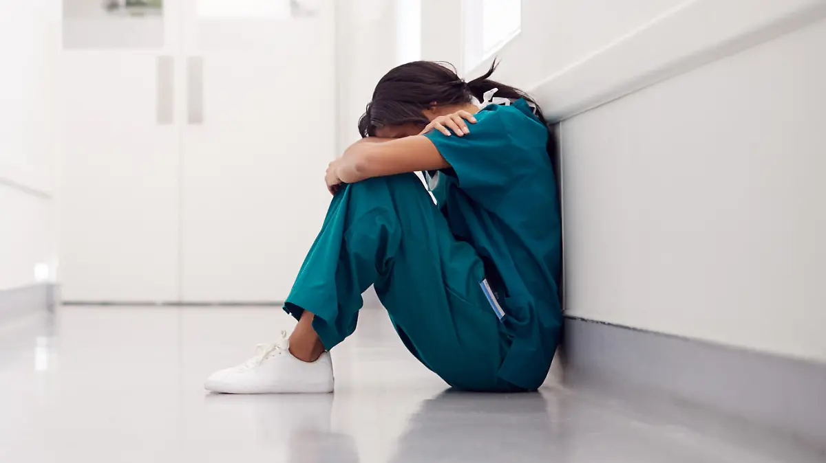 Stressed And Overworked Female Doctor Wearing Scrubs Sitting On Floor In Hospital Corridor