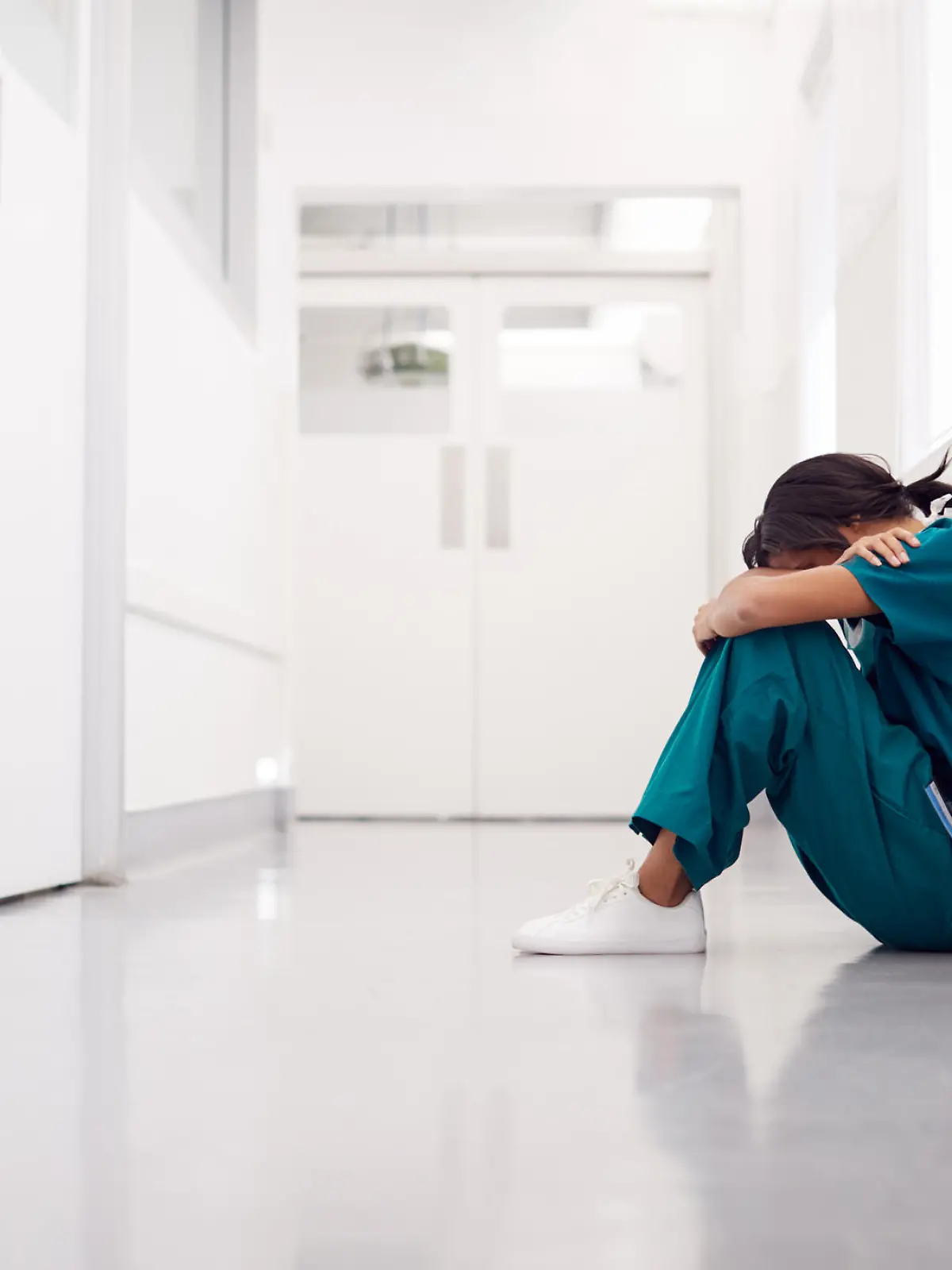 Stressed And Overworked Female Doctor Wearing Scrubs Sitting On Floor In Hospital Corridor