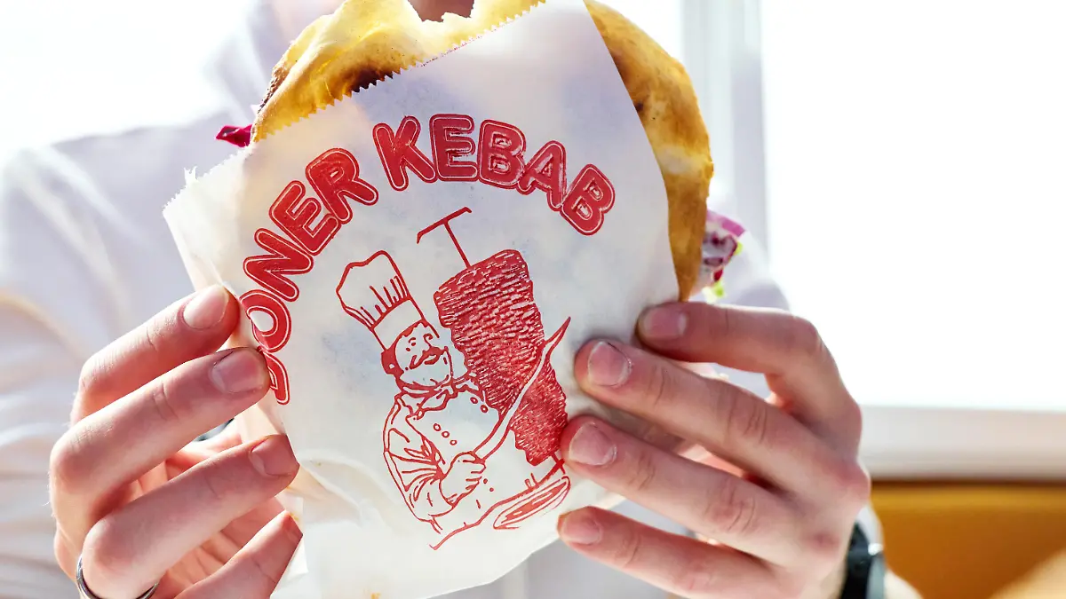 Mann hält einen Döner Kebab in einer bedruckten Dönerverpackung. Fastfoodgericht im Restaurant oder Imbiss. Typisches Streetfood mit Fleisch, Salat und Sauce