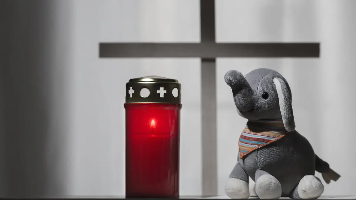 Grabkerze neben Stofftier mit einem Kreuz im Hintergrund.