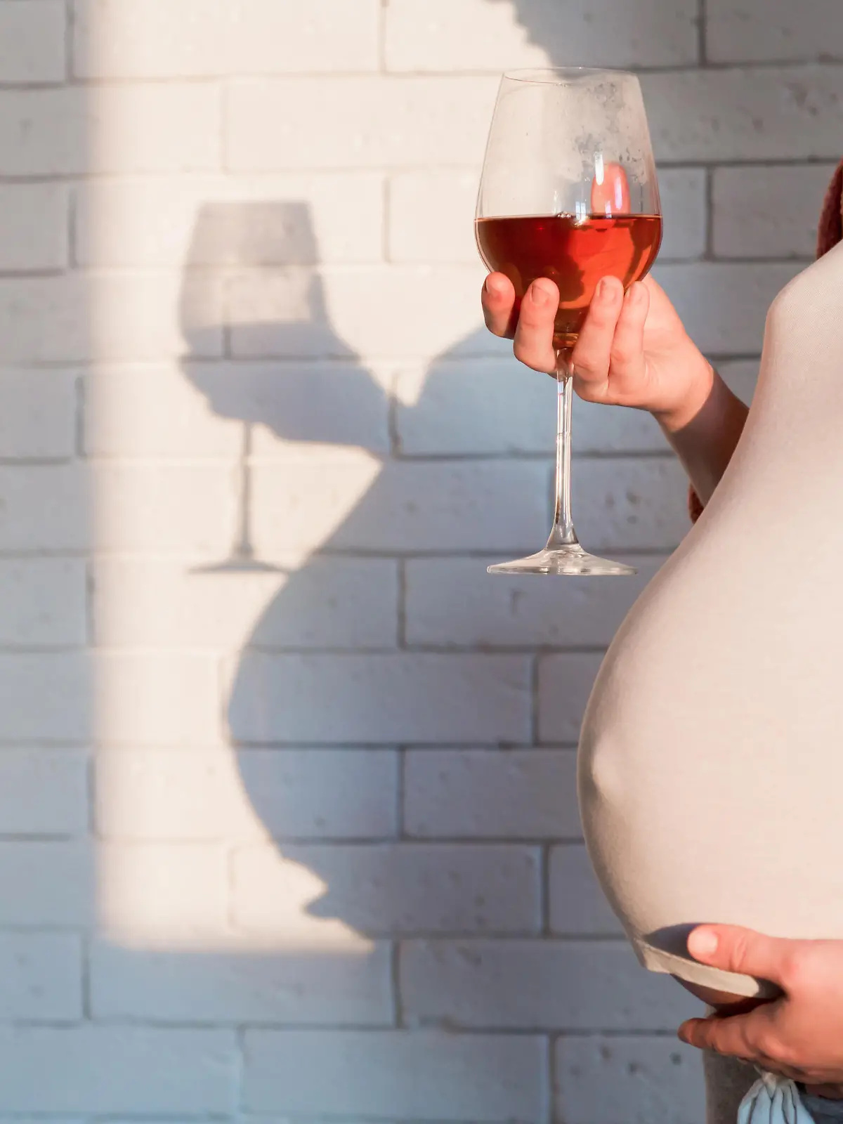 Schwangere Frau hält ein Glas Rotwein in der einen Hand und hält mit der anderen Hand ihren Babybauch fest