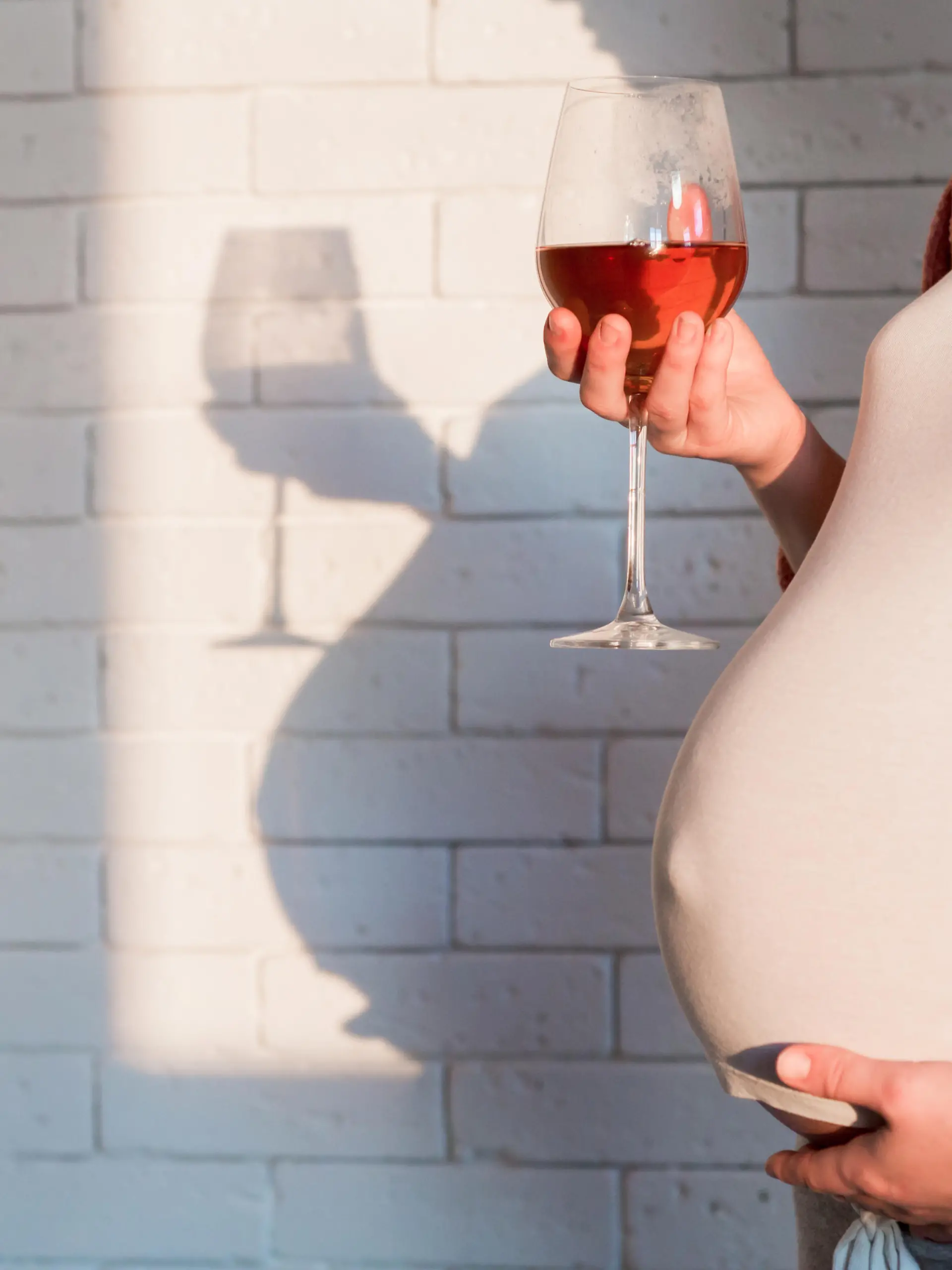 Schwangere Frau hält ein Glas Rotwein in der einen Hand und hält mit der anderen Hand ihren Babybauch fest