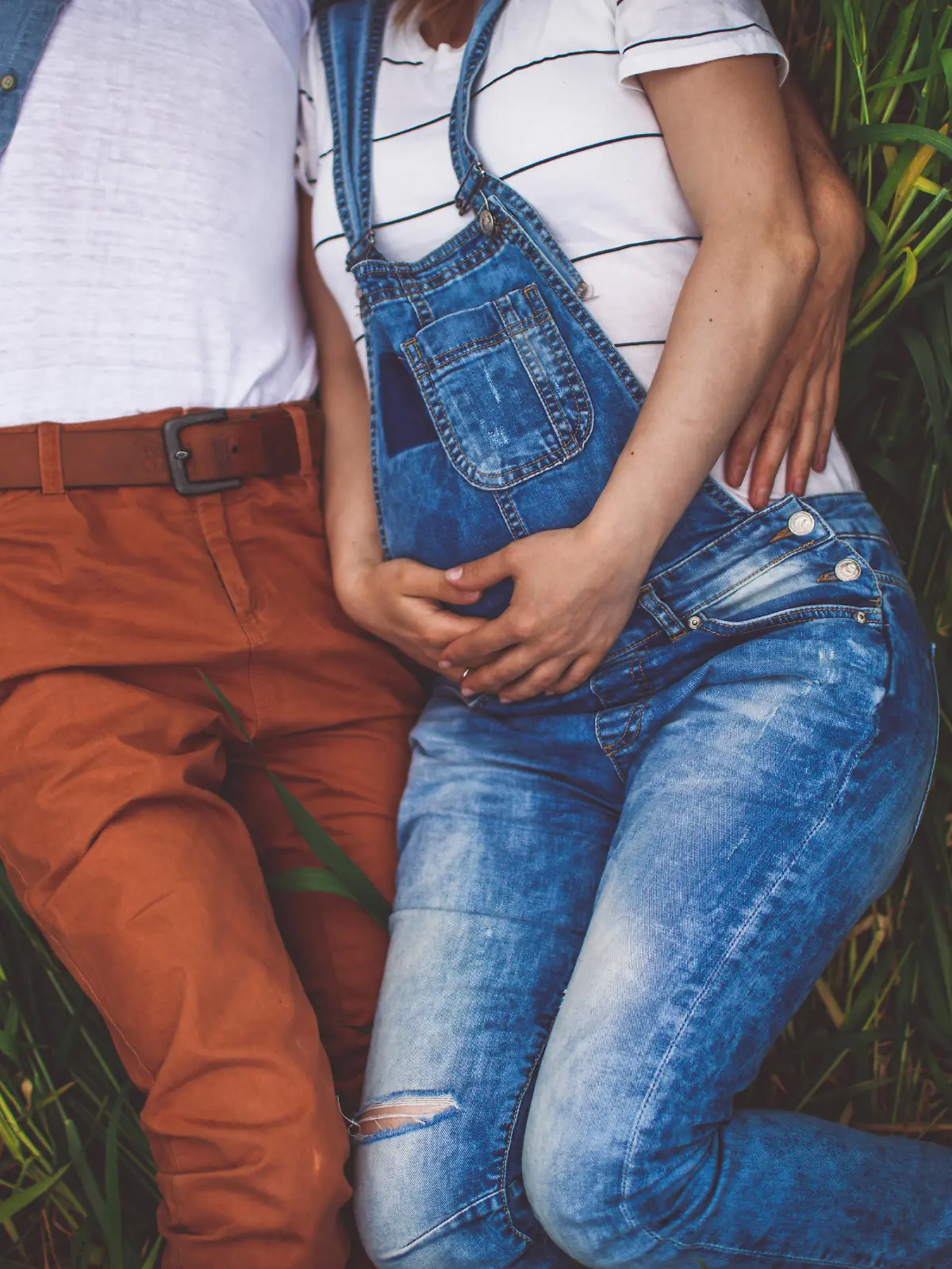 Photo of a young couple, soon to become parents, resting in the middle of the field on a beautiful spring day while expecting a new life to begin. Baby bump is in a focus, while parents are out of the frame