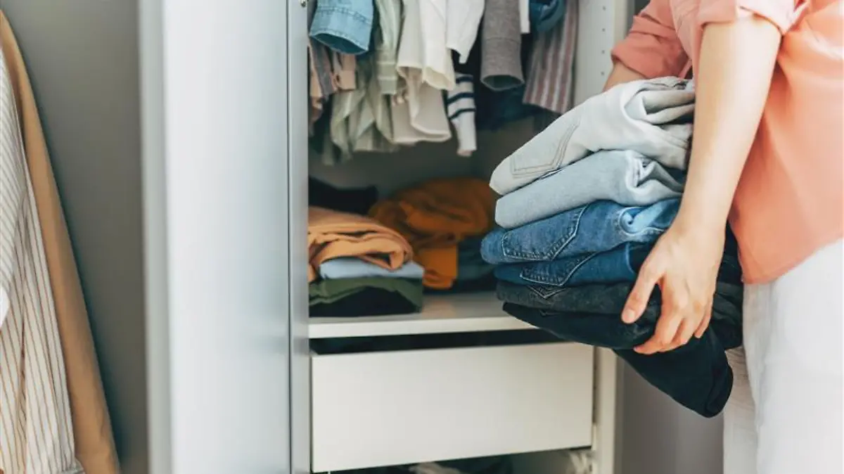 Frau hält einen Stapel Jeans vor einem Kleiderschrank.