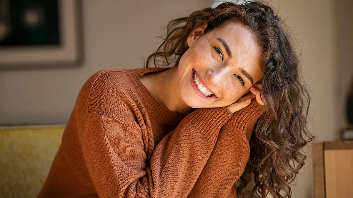 Happy young woman sitting on sofa at home and looking at camera. Portrait of comfortable woman in winter clothes relaxing on armchair. Portrait of beautiful girl smiling and relaxing during autumn.