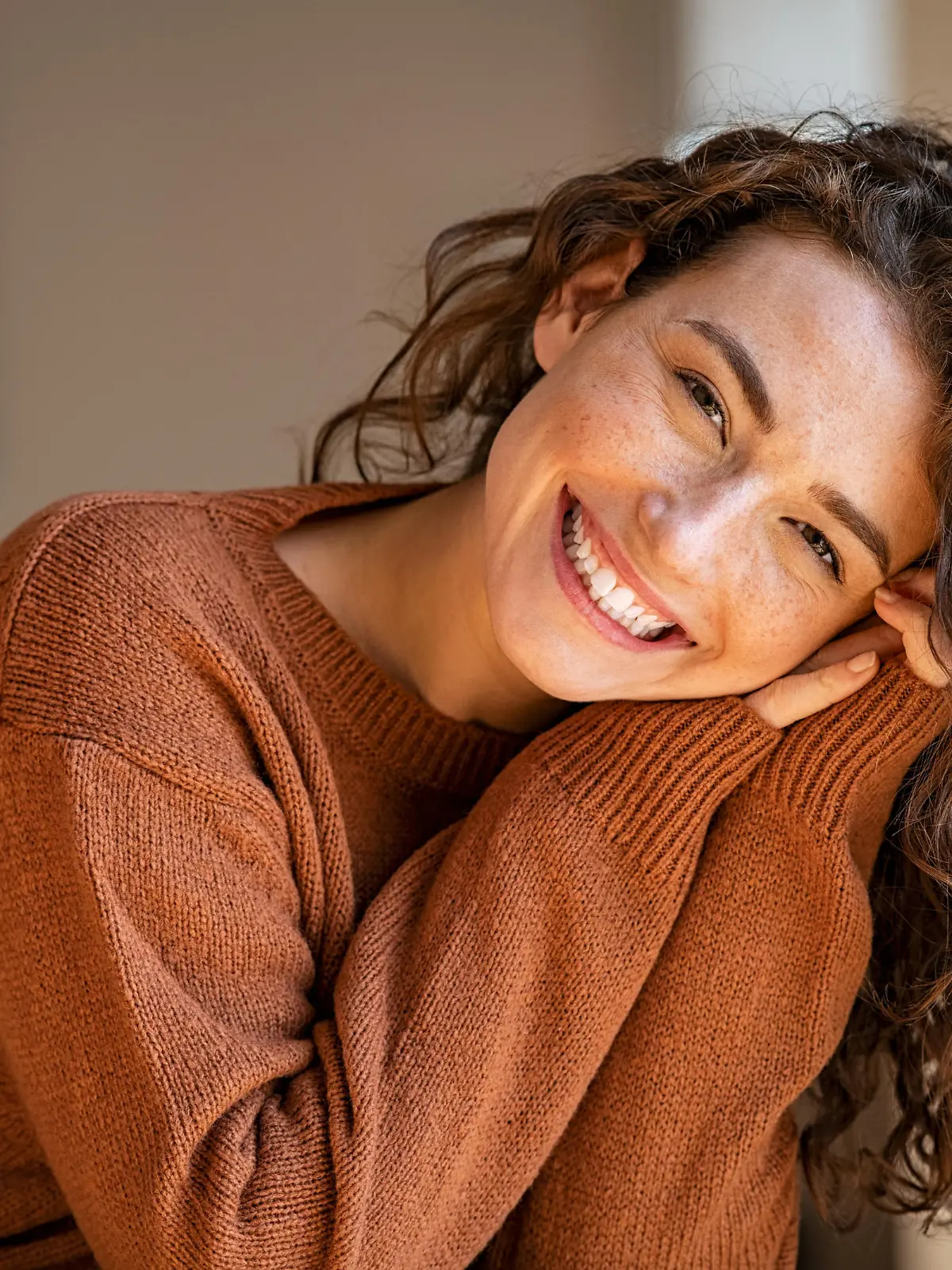 Happy young woman sitting on sofa at home and looking at camera. Portrait of comfortable woman in winter clothes relaxing on armchair. Portrait of beautiful girl smiling and relaxing during autumn.