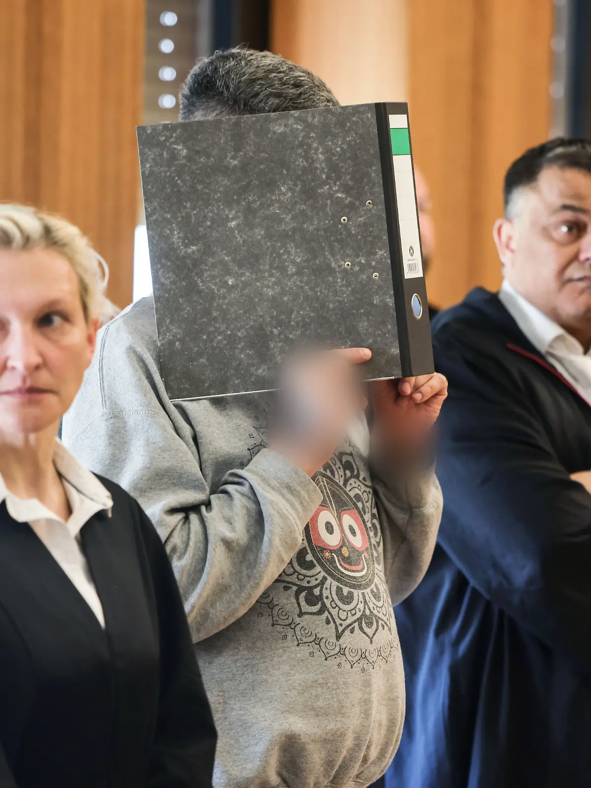 Der angeklagte Krankenpfleger hält sich zwischen seinen Anwälten einen Aktenordner vor das Gesicht. 