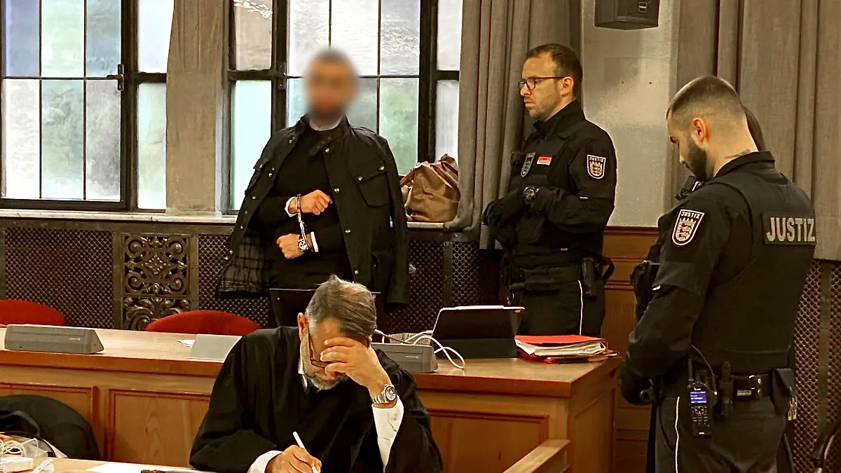 Der Angeklagte (hinten links) steht in Handschellen im Verhandlungssaal im Landgericht neben Justizvollzugsbeamten. 