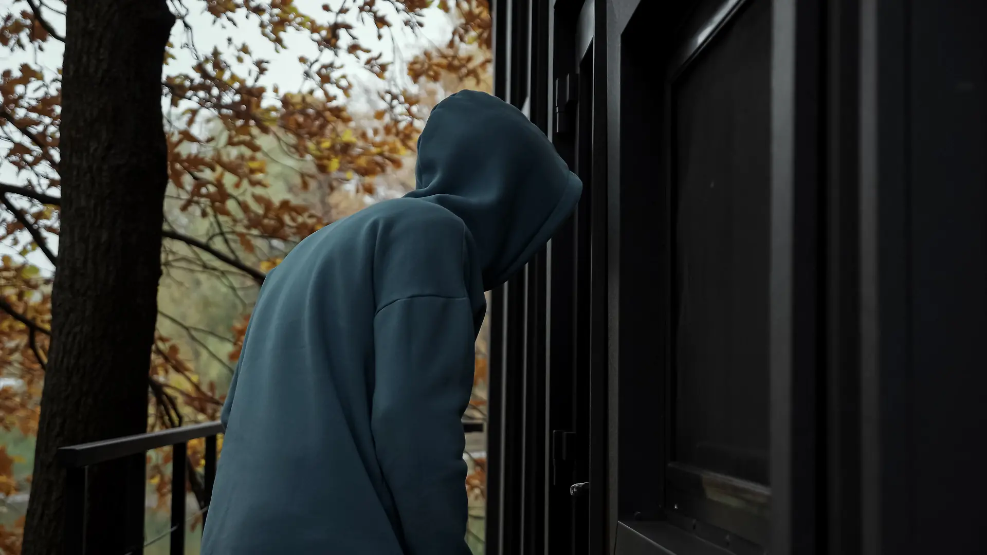 Ein Mann in dunkler Kleidung und Kapuze, der im Herbst um ein Haus herumschleicht und nach einer Möglichkeit des Einbruchs sucht.