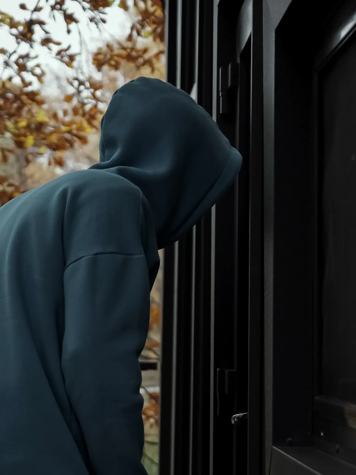 Ein Mann in dunkler Kleidung und Kapuze, der im Herbst um ein Haus herumschleicht und nach einer Möglichkeit des Einbruchs sucht.