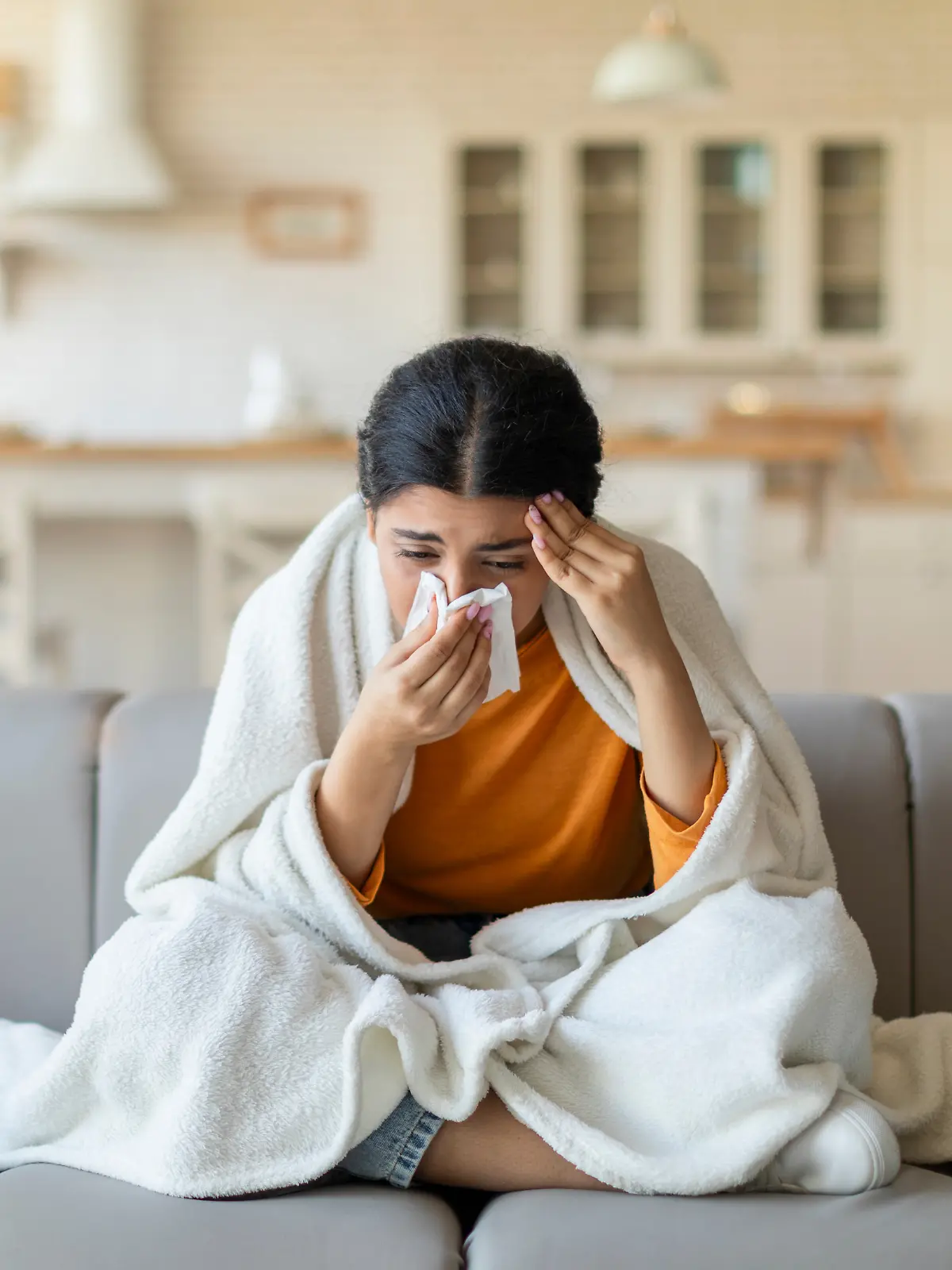 Erkältete Frau sitzt in einer Decke eingewickelt auf der Couch und schnäuzt sich die Nase.