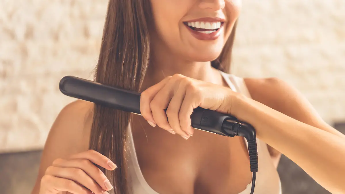Cropped image of beautiful young woman in white undershirt using a hair straightener and smiling while looking into the mirror in bathroom