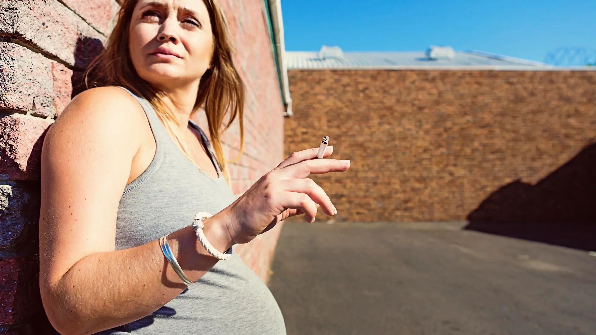 A pregnant woman stands in a parking lot as she guiltily sneaks a cigarette, knowing it's bad for her and her baby. Copy space on the tarmac.