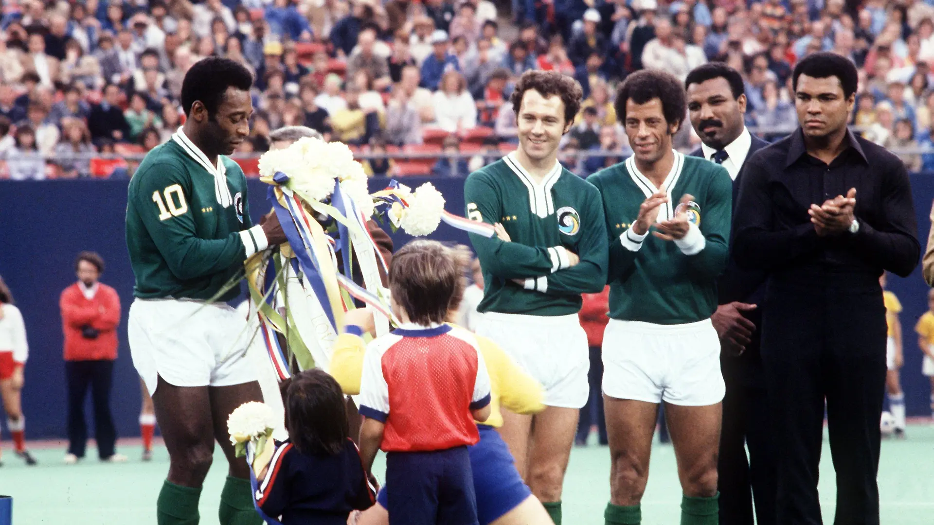 Der brasilianische Fußball-Star Pele (l) erhält am 1.10.1977 im New Yorker Giants-Stadion vor seinem letzten Spiel für Cosmos New York Blumen aus Kinderhänden. Er wird flankiert von seinen beifallspendenden Mitspielern Franz Beckenbauer (4.v.r), seinem Landsmann Carlos Alberto (3.v.r) und der US-Box-Legende Muhammad Ali (r). Pele beendet mit diesem Spiel seine einzigartige Karriere. Er spielte im letzten Teil seiner Laufbahn zwei Jahre und vier Monate für Cosmos New York, bestritt 108 Spiele und schoß 68 Tore. An diesem Tag schießt er sein letztes von insgesamt 1282 Toren (in 1364 Spielen).