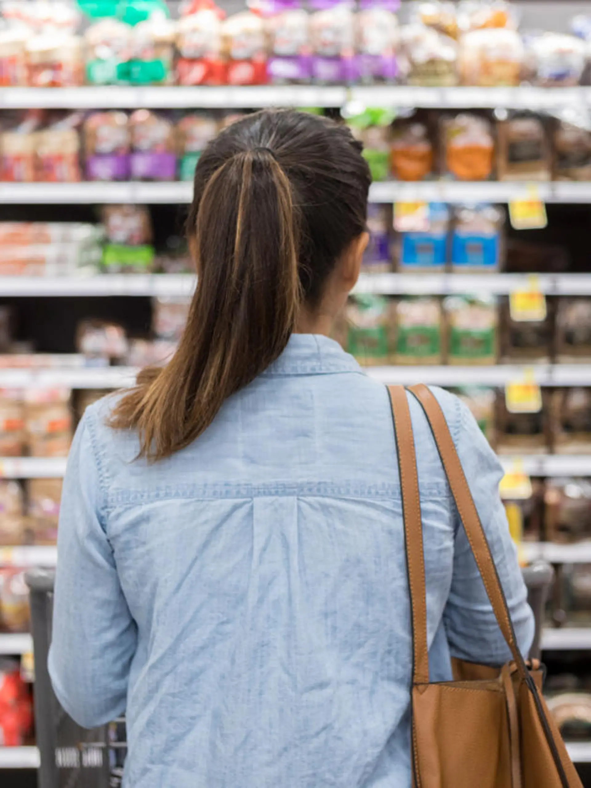 In this rear view, an unrecognizable woman stands with a shopping cart in front of a shelf full of food in the bread aisle of a grocery store.