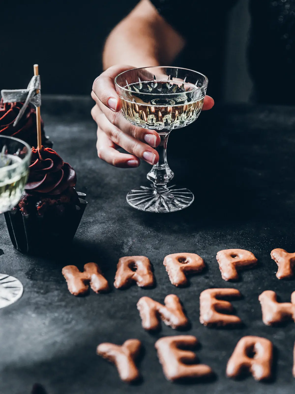 Happy new year cookie on table with cupcakes and a woman sitting holding a glass of champagne.