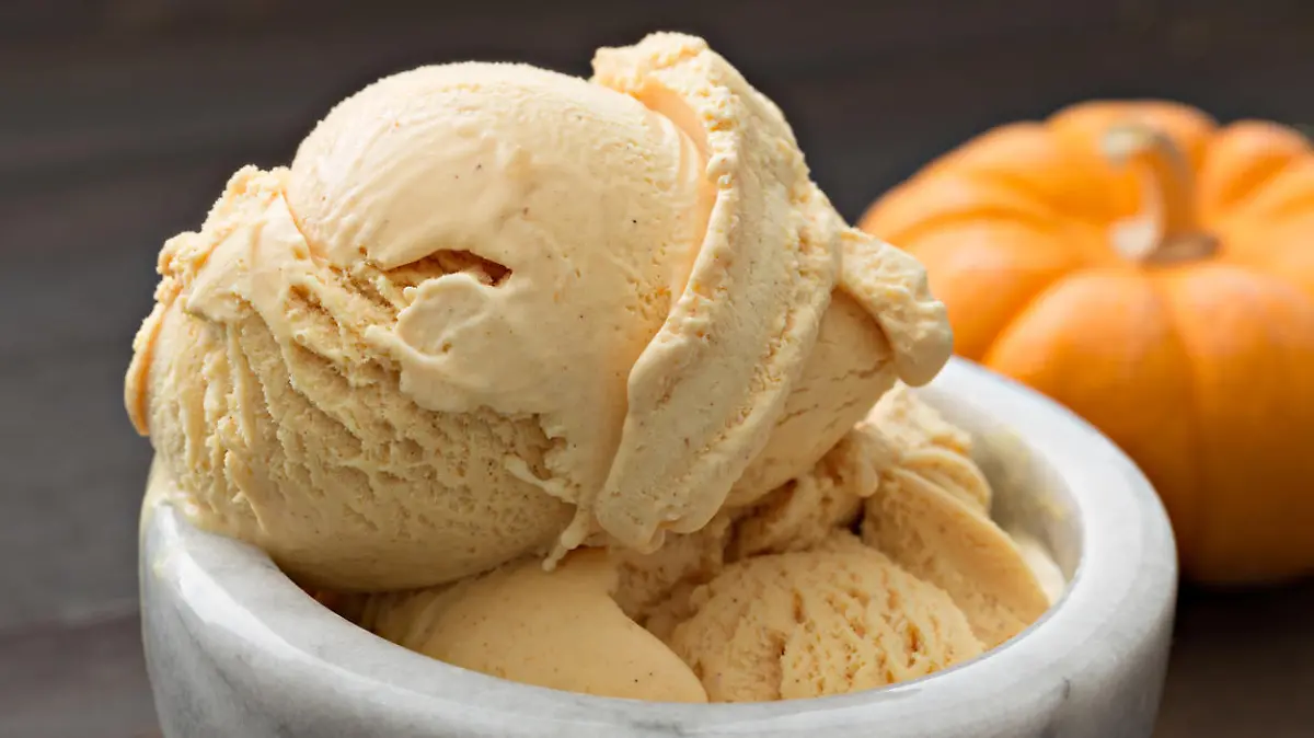 A close up, horizontal photograph of a scoop of pumpkin ice cream in a bowl, a small pumpkin is sitting next to the dessert.