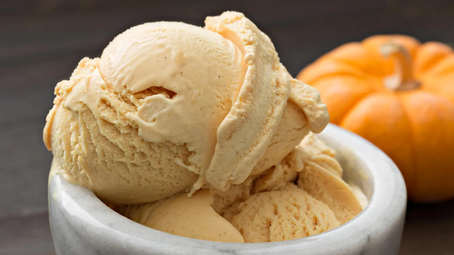 A close up, horizontal photograph of a scoop of pumpkin ice cream in a bowl, a small pumpkin is sitting next to the dessert.