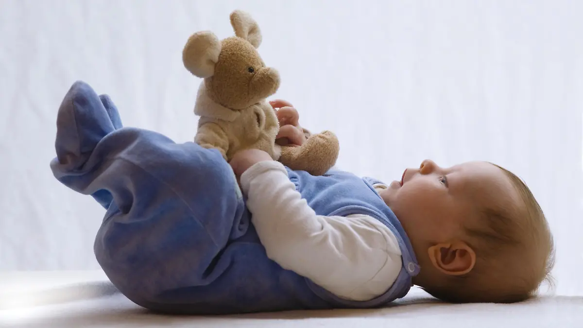 Baby liegt im Strampler auf dem Bett und spielt mit Stofftier